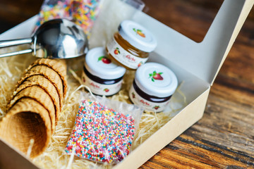 A Kit For Making Ice Cream: Spoon For Ice Cream, Ice Cream Cones, Jam, Topping, Chocolate, Sprinkles, Coconut. In Carton Wooden Natural Background. The Concept Set Surprise, Gift Idea