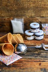 A Kit For Making Ice Cream: Spoon For Ice Cream, Ice Cream Cones, Jam, Topping, Chocolate, Sprinkles, Coconut Shavings. On Wooden Background. The Concept Of A Set, Surprise, Gift Idea