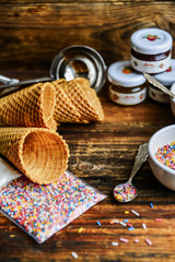 A Kit For Making Ice Cream: Spoon For Ice Cream, Ice Cream Cones, Jam, Topping, Chocolate, Sprinkles, Coconut Shavings. On Wooden Background. The Concept Of A Set, Surprise, Gift Idea