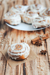 Freshly baked warm iced cinnamon buns on baked paper. Sweet homemade pastry baking on with cup of coffee and spices on wooden background.