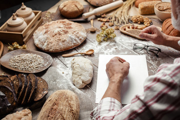 Fototapeta premium Portrait of smiling Baker's hands chef writing a new recipe of homemade bread on white paper shift, surrounded by ruralorganic bread variety