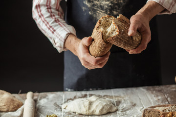 Man breaking off piece of rustic rye bread, standing at table with dough in flour, closeup