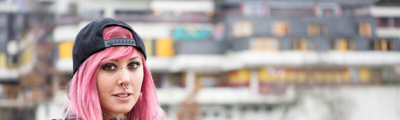 young alternative woman with piercings and pink hair in front of defocused housing estate in the...