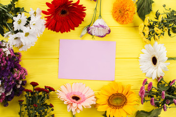 Variety of multicolored blossoming flowers and blank paper. Yellow wooden desk background.