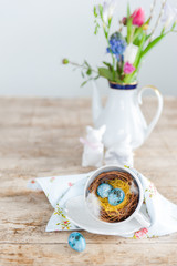 Bright speckled easter eggs in a nest from a cup in the foreground. Porcelain hares and a vase with flowers in the background