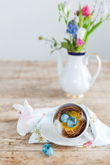 Bright beautiful speckled easter eggs in a nest from a cup in the foreground. Porcelain hares and a vase with flowers in the background