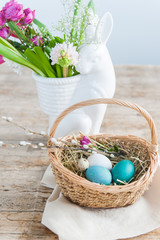 Wicker basket with Easter eggs in the foreground and a rabbit vase with spring flowers in the background.