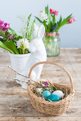 Wicker basket with Easter eggs in the foreground and a white rabbit vase with spring flowers in the background.