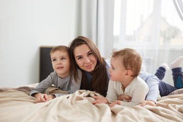 Happy family. Young mother dressed in light blue pajama lays with her two little sons on the bed with beige blanket in the bedroom with big window