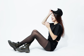 Smiling young dark-haired girl dressed in a black top, shorts, tights and cap poses sitting on the floor on the white background in the studio