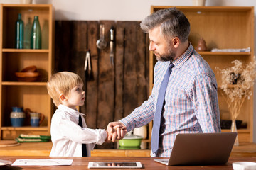 Serious confident handsome bearded father in shirt and tie standing in kitchen and making handshake with little son in formal shirt