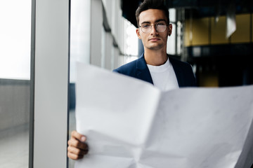 Fototapeta premium Architect in stylish clothes holds sheet with drawing in his hand and talks by phone on the background of a modern glass multistory building