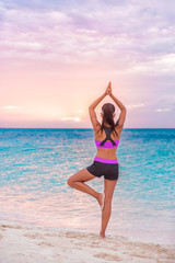 Yoga wellness class on beach at sunset. Girl practicing meditation standing on one leg training balance barefoot on sand enjoying sun and ocean view.