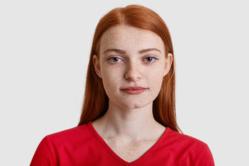 Headshot of attractive red haired European woman with freckled skin, looks seriously at camera, has minimal make up, wears red sweater, isolated over white background. Natural beauty concept - obrazy, fototapety, plakaty