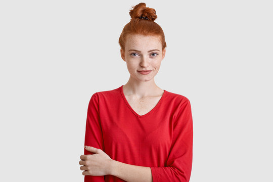 Photo of attractive woman with red hair, keeps hand over chest, wears casual clothes, has freckled skin, isolated over white background. Pretty youngster models indoor. People and beauty concept