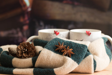Two stars of aniseed seeds spice are on beautiful warm knitted striped scarf with background of two mugs with red hearts and burning wooden logs in fireplace. Concept: Valentine's Day holiday card.