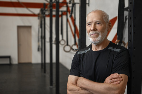 Picture of handsome muscular European male pensioner with white beard and blue eyes relaxing in gym after heavy exercises workout, keeping arms crossed on his chest and smiling confidently