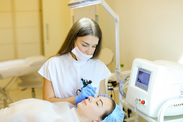 Young cosmetologist making permanent makeup for girl at beauty salon.