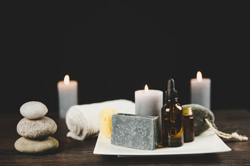 Men`s spa relaxation concept. Different day spa products( soap bar, beard oil, candles burning, rolled towel) on white ceramic tray on wooden table, dark black background.