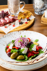 Greek salad with feta cheese, olives and herbs, served in a white plate on a table in a restaurant.
