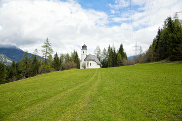 View of a small white church on a hill in the middle of a vibrant green alpine meadow in Ehrwald, Austria.