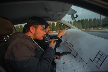 Two robbers sitting inside a car with guns looking at the blueprint of the building they are about to rob.