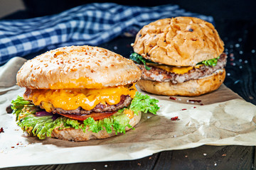Two burgers with meat, spices and sauce on a craft paper on a black wooden background. A picture for advertising and recipes. Close up view on American Fast Food. Copy free space for logo, text.