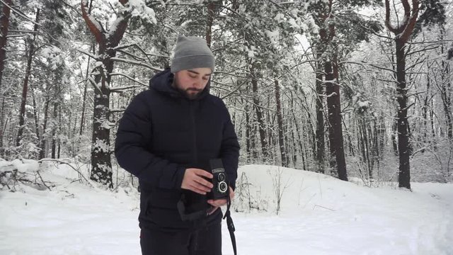 Photographer is standing in the deep snow in the winter forest, framing and imagining a shot in his mind befor taking a shot on his vintage medium film camera. Front view.
