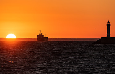 Hazy ocean sunset with ship and lighthouse silhouettes.