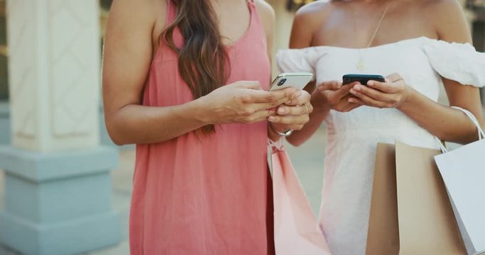 Two beautiful young woman texting on their phones with shopping bags on arms