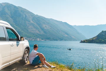 young man sitting near suv car at seaside with beautiful view of sea bay with mountains