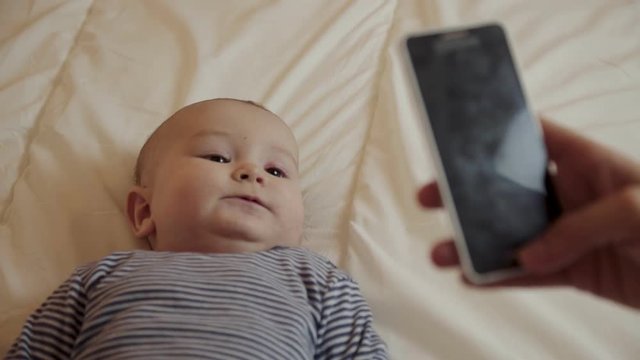 Mother with smartphone taking pictures of cute baby. Cropped view of parent using cell phone and photographing adorable infant lying on bed. Technology concept
