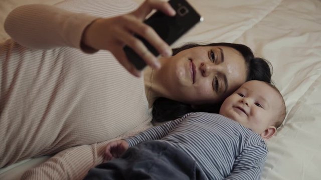 Happy mother using smartphone with baby on bed. Smiling young woman holding cell phone and lying with adorable infant kid on bed. Parenthood concept