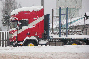 Red truck with semi-trailer platform stands in the snow on parking near warehouse on roadside - bad weather, loading, rest of the driver, side view
