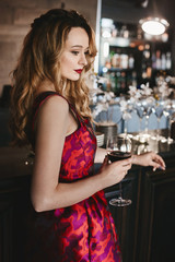 Fashionable blonde model girl with bright red lips and with natural makeup in the red-pink dress stands at the bar with the glass of red wine in her hand and posing in luxury dark interior