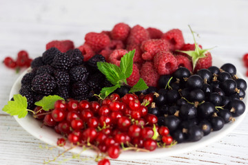 Black and red currants, black and red raspberries on a plate with a leaf of raspberry, white