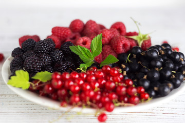 Black and red currants, black and red raspberries on a plate with a leaf of raspberry, white