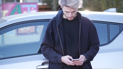Handsome young man using mobile phone to type a message or surfing the internet, while leaning on the side of a car - Powered by Adobe