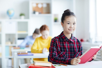 Little schoolgirl with open book siting by desk in classroom during lesson of literature