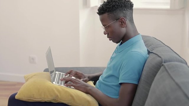 Young african-american  freelancer  using laptop. Man with computer on pillow working on cozy sofa. Cozy workplace concept.