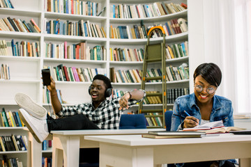 Beautiful African-American students work in the library. The girl smiles and working with the laptop and, reads the books and the boy dances while listening to music.