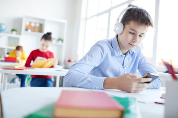 Restful schoolboy in headphones choosing song from playlist in smartphone at break between lessons