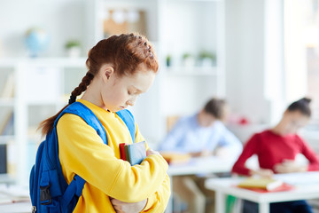 Fototapeta premium Sad or offended schoolgirl with backpack and books standing in classroom among classmates