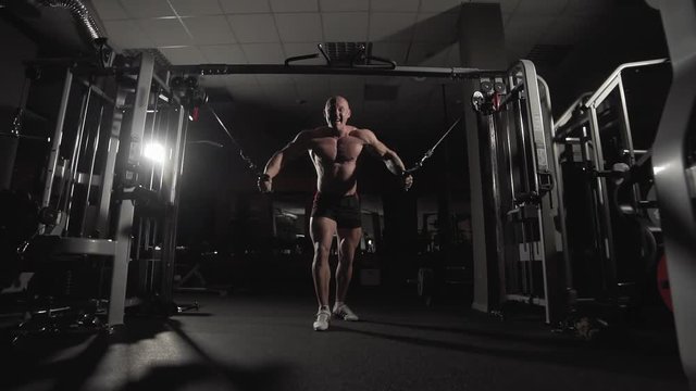 Male bodybuilder training chest muscles exercise using cable cross over machine for body mass. Workout and bodybuilding sport concept. Man in dark gym doing pulls weight exercise in slow motion.