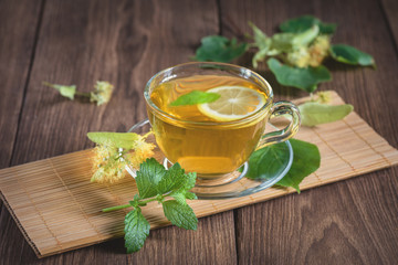 A cup of medicinal, lime tea, standing on a wooden table, surrounded by fragrant linden flowers, in the rays of sunlight.