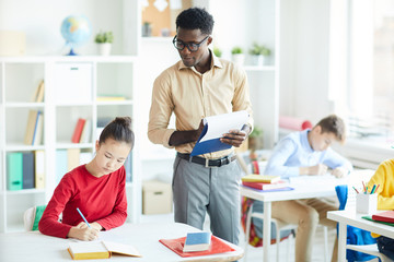 Young confident teacher making notes in document while standing by desk of diligent pupil at lesson