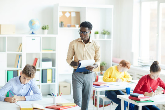Group of diligent schoolkids carrying out written assignment while young teacher walking along rows at lesson