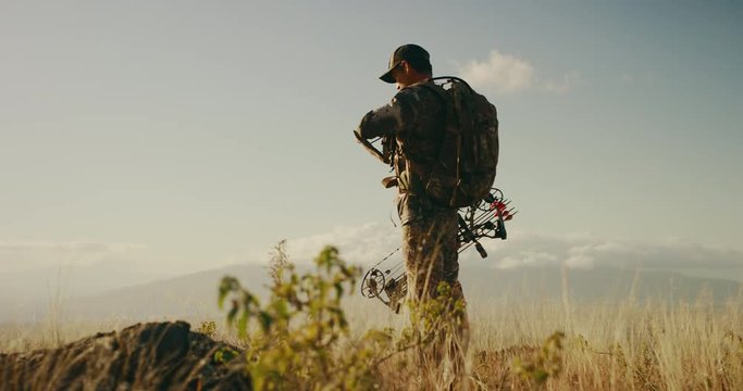 Orbit shot of majestic bowhunter on grassy knoll raising his binoculars