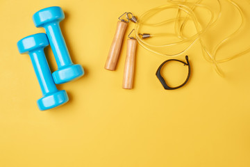 Flat lay composition of blue dumbbells, jump rope and fitness tracker on a yellow background with copy space