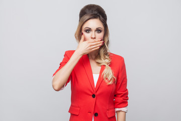 Portrait of shocked beautiful business lady with hairstyle and makeup in red fancy blazer, standing and covering her mouth and looking at camera with big eyes. studio shot, isolated on grey background